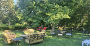 Linden Historic Bed & Breakfast backyard garden with wrought-iron patio seating, cushioned chairs, blooming potted flowers, and shaded lawn surrounded by mature trees in Natchez, Mississippi.