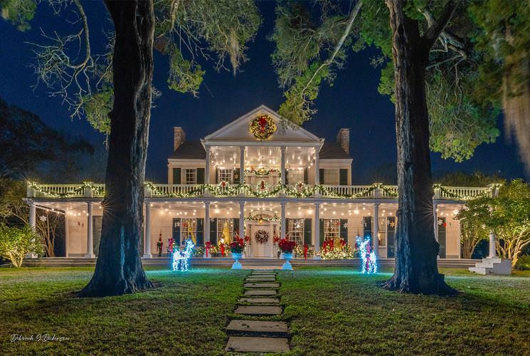 Exterior view of the property painted white with dark shutters, front porch, and second-level balcony all surrounded by green grass and large trees all with Christmas decorations at night
