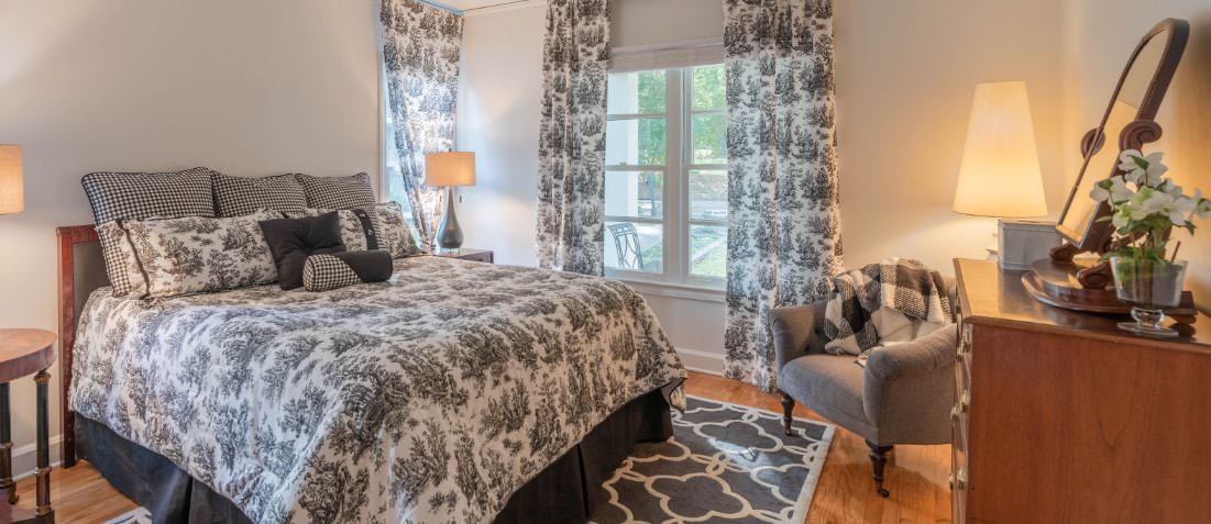 Queen bed with black and white comforter with matching drapes on windows. Stands with lamps and antique dresser with mirror.