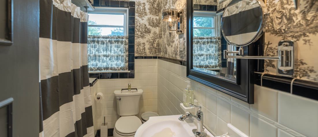Slender bathroom with shower on left with black and white striped curtain. Sink with mirror and white tiled walls. Window with valance over toilet.