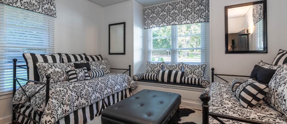 Sunroom with 2 daybeds with black and white spreads separated by leather topped ottoman. Windows with valances and mini blinds.